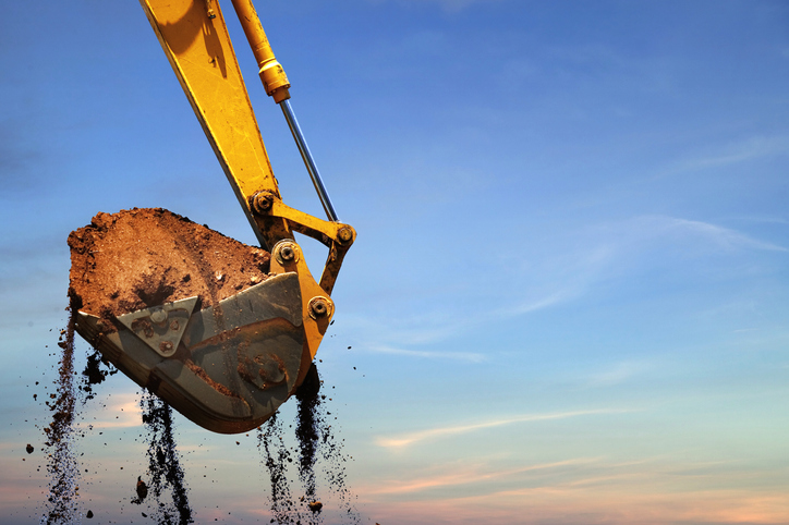 A yellow excavator bucket lifting soil against a clear blue sky, symbolizing excavation work and land preparation.